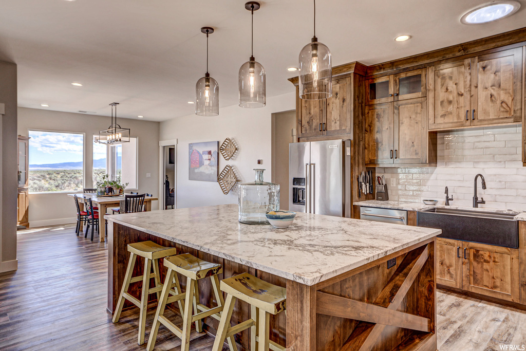 Kitchen with decorative light fixtures, backsplash, light hardwood floors, a kitchen island, appliances with stainless steel finishes, a center island with sink, and light stone counters
