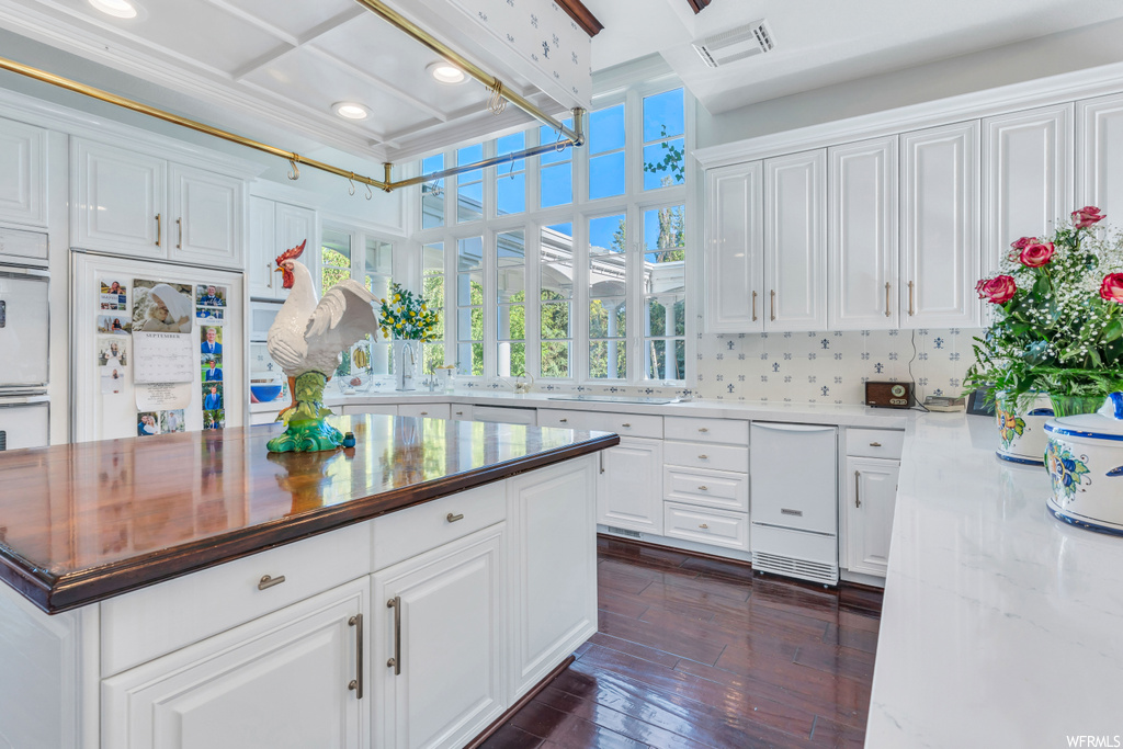 Kitchen featuring coffered ceiling, white cabinets, white oven, dark hardwood flooring, light stone countertops, backsplash, and built in fridge