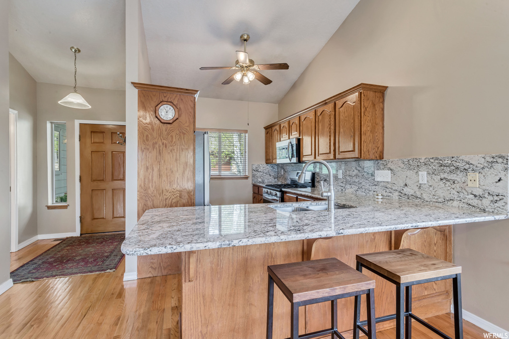 Kitchen with appliances with stainless steel finishes, a center island, light stone countertops, brown cabinets, vaulted ceiling, hanging light fixtures, light hardwood flooring, and backsplash