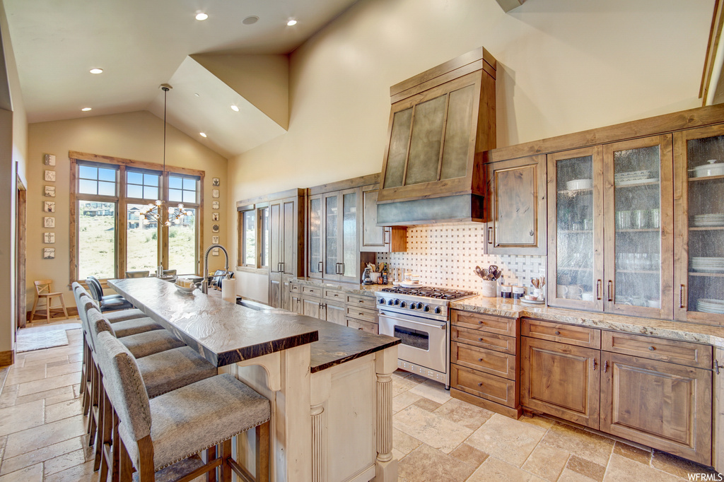 Kitchen with a kitchen island, lofted ceiling, light stone countertops, brown cabinets, high end stainless steel range, premium range hood, backsplash, a high ceiling, and light tile floors
