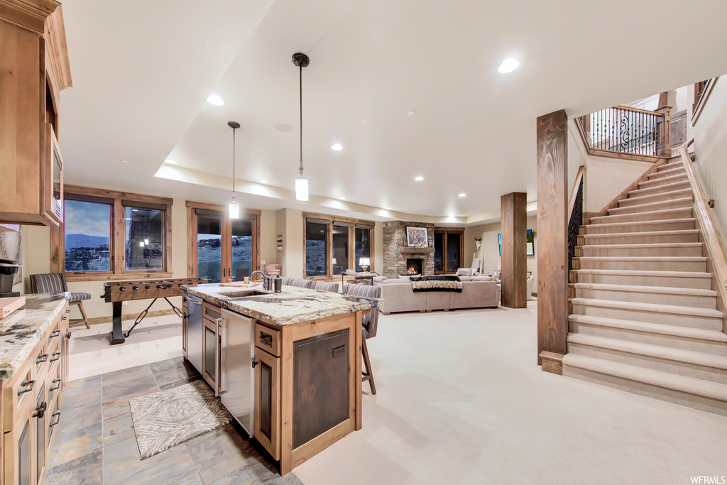Kitchen featuring stainless steel dishwasher, pendant lighting, a raised ceiling, a fireplace, light stone countertops, a center island with sink, and light tile floors