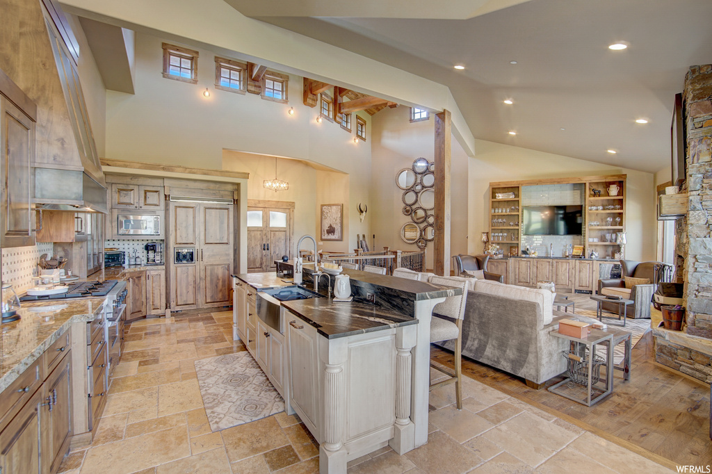 Kitchen featuring appliances with stainless steel finishes, custom range hood, light tile flooring, light stone countertops, vaulted ceiling, kitchen island with sink, backsplash, and a high ceiling
