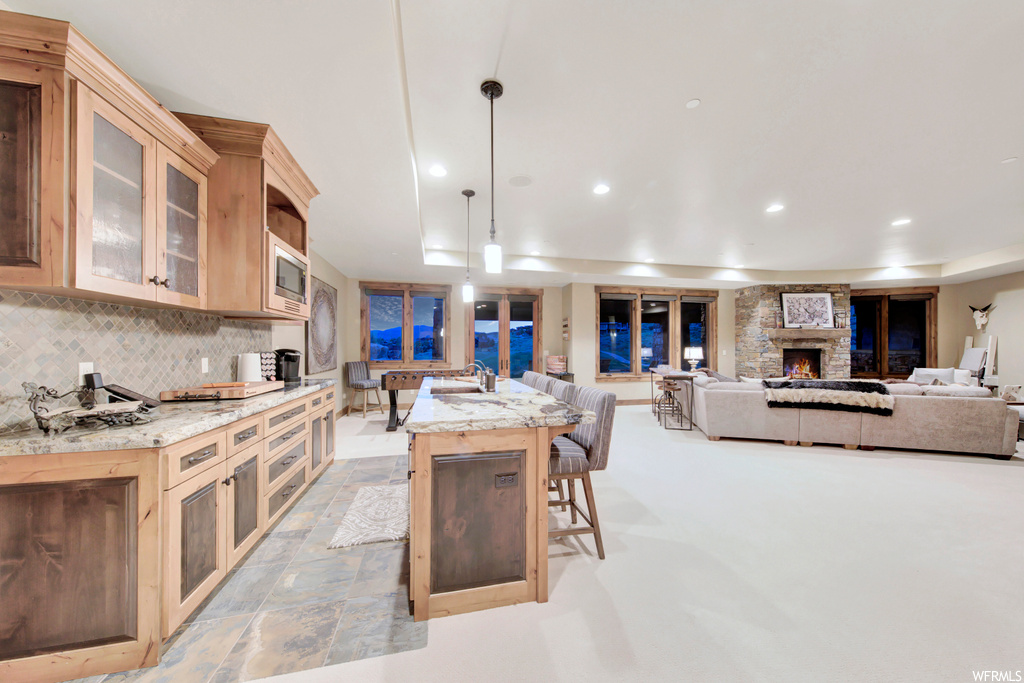 Kitchen featuring backsplash, a kitchen island, light tile floors, pendant lighting, a fireplace, stainless steel microwave, and light stone counters