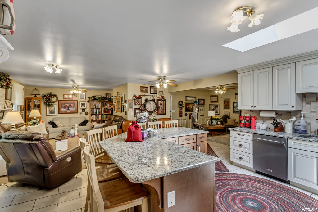 Kitchen featuring stainless steel dishwasher, backsplash, white cabinets, a kitchen island, light tile floors, ceiling fan, light stone countertops, and a skylight