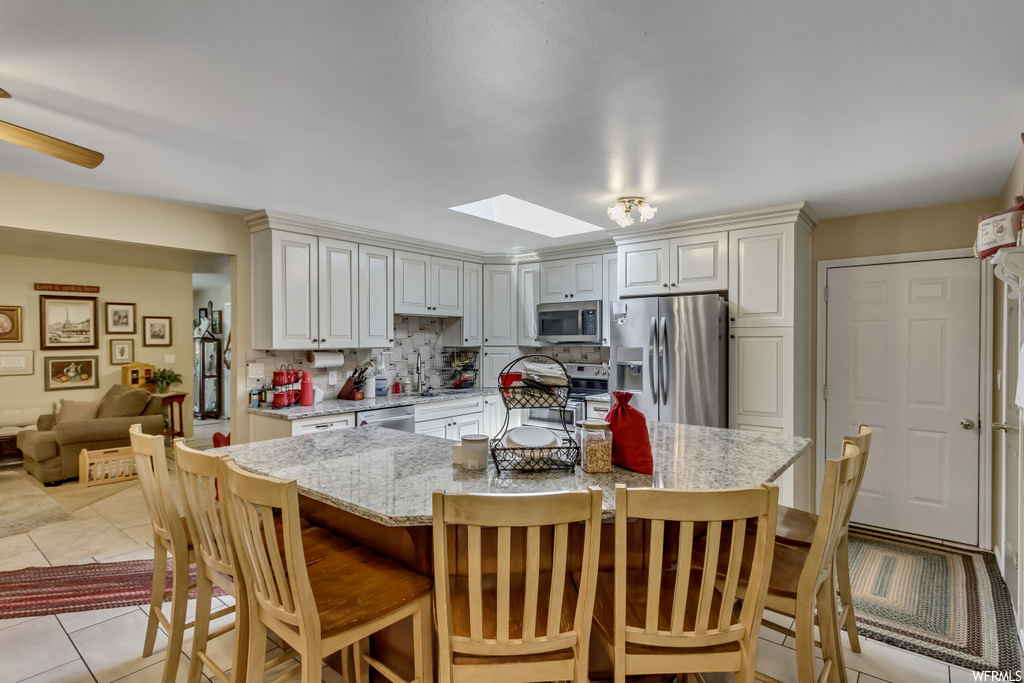 Kitchen with backsplash, white cabinetry, a center island, light tile flooring, stainless steel appliances, a skylight, and light stone counters