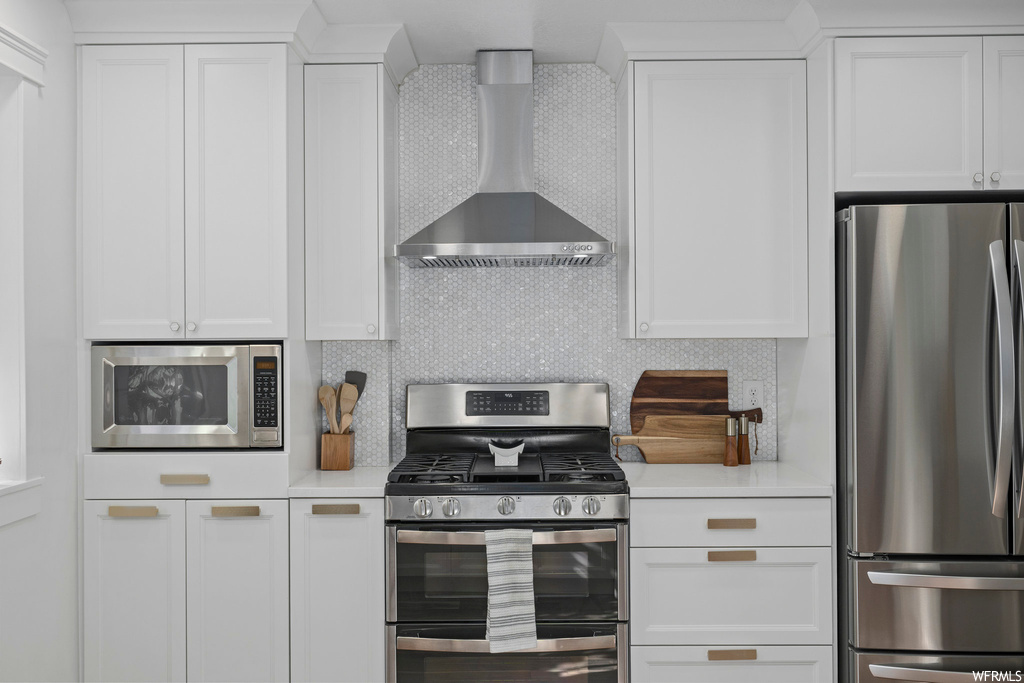 Kitchen featuring backsplash, white cabinets, wall chimney range hood, light countertops, and stainless steel appliances