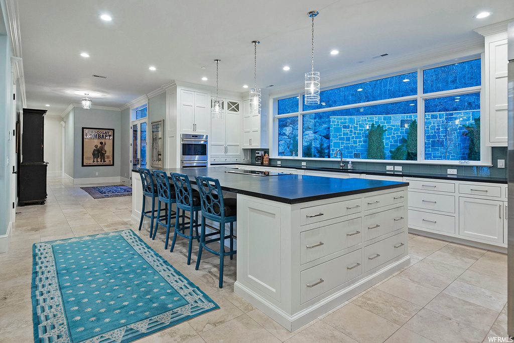 Kitchen featuring ornamental molding, hanging light fixtures, backsplash, white cabinetry, a center island with sink, light tile floors, stainless steel double oven, and dark countertops