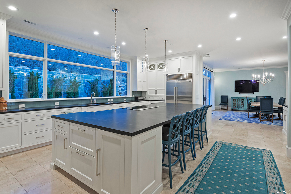 Kitchen featuring stainless steel built in refrigerator, a center island, crown molding, a kitchen island with sink, dark countertops, light tile flooring, white cabinets, hanging light fixtures, and backsplash