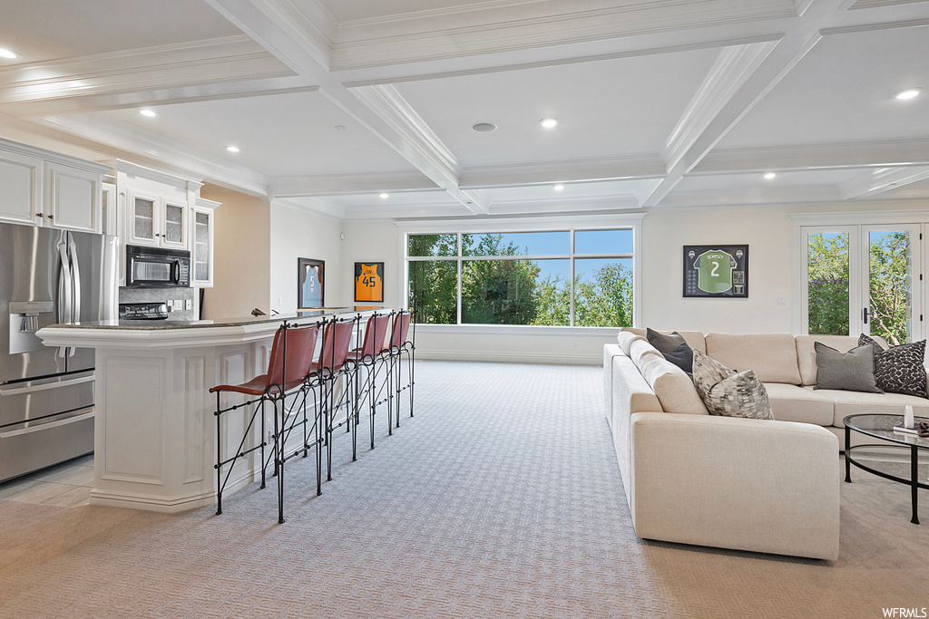 Interior space with beamed ceiling, white cabinetry, a center island, coffered ceiling, a wealth of natural light, light carpet, light countertops, ornamental molding, and stainless steel fridge with ice dispenser