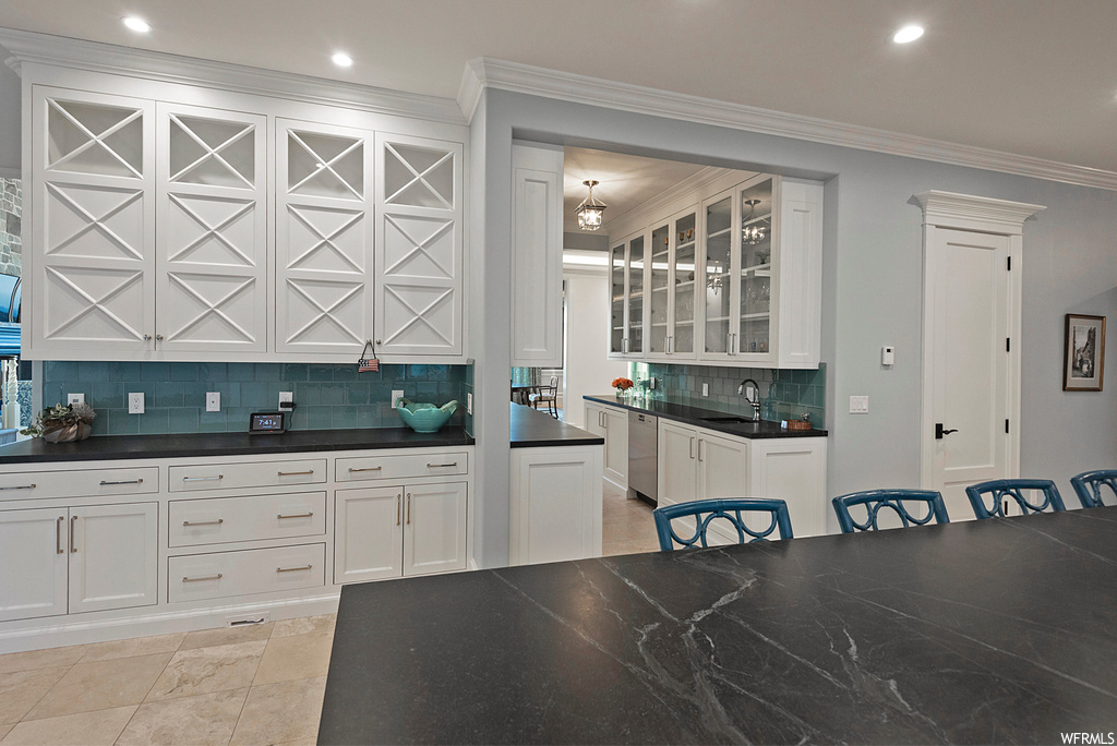 Kitchen with stainless steel dishwasher, backsplash, white cabinetry, crown molding, tile floors, and dark countertops