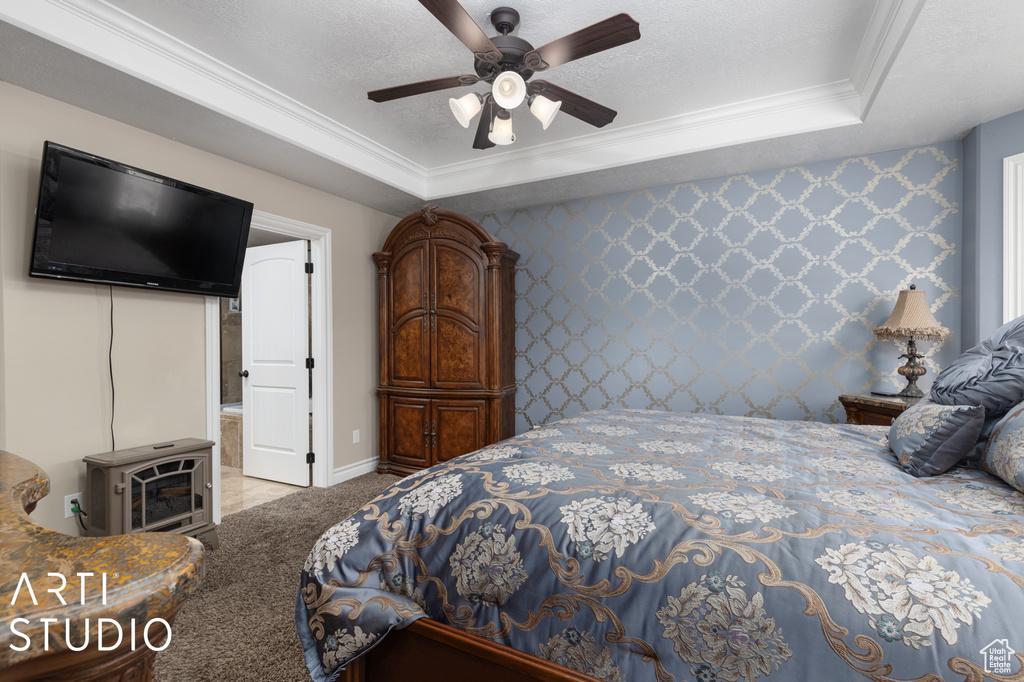 Carpeted bedroom with ceiling fan, a wood stove, a tray ceiling, connected bathroom, and ornamental molding