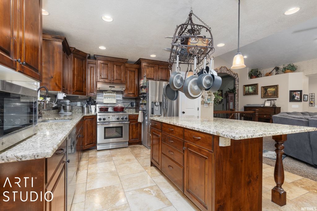 Kitchen with appliances with stainless steel finishes, light tile flooring, light stone counters, dark brown cabinets, hanging light fixtures, and backsplash