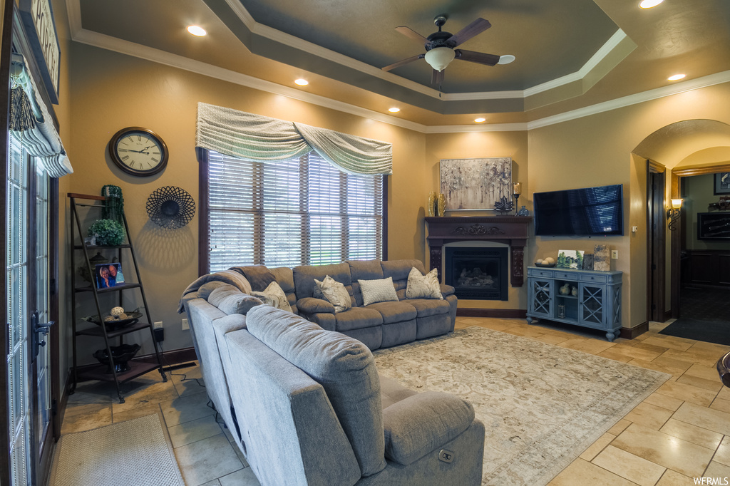 Living room featuring crown molding, light tile flooring, a raised ceiling, a fireplace, and ceiling fan