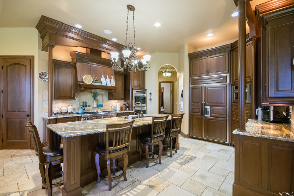 Kitchen with a kitchen island, custom range hood, stainless steel double oven, light tile flooring, dark brown cabinetry, light stone counters, paneled built in fridge, and backsplash