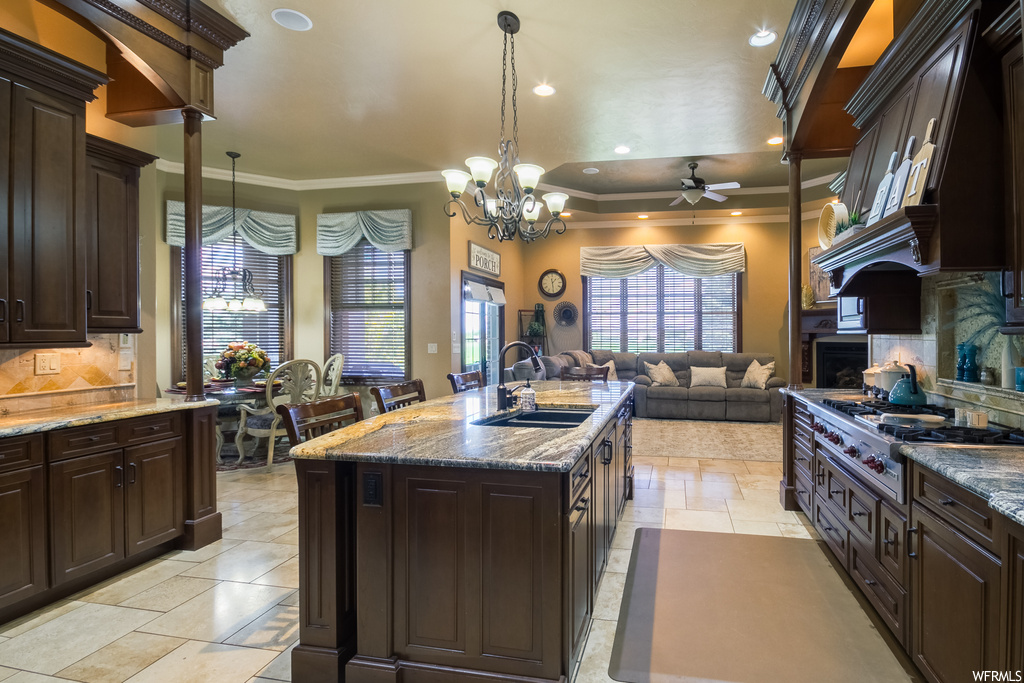 Kitchen with stainless steel gas cooktop, kitchen island with sink, backsplash, custom exhaust hood, light tile floors, dark brown cabinetry, ceiling fan, crown molding, and light stone counters