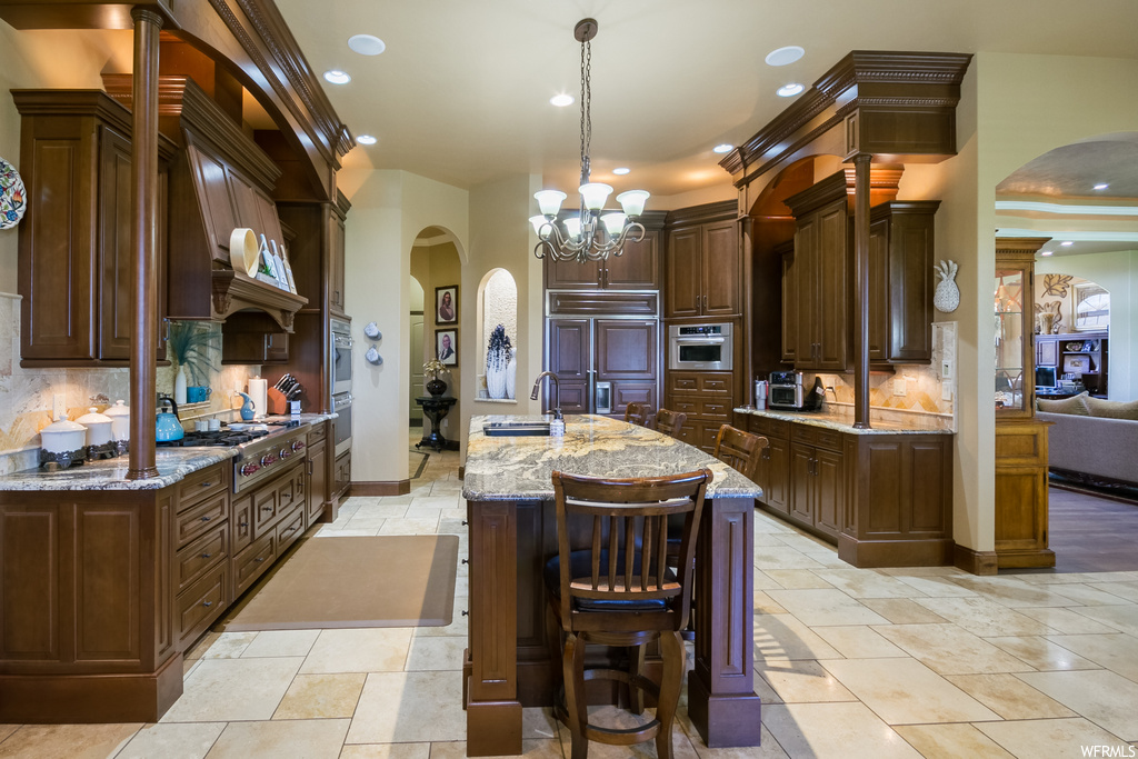 Kitchen with kitchen island with sink, backsplash, a notable chandelier, dark brown cabinetry, light stone countertops, stainless steel appliances, and light tile flooring