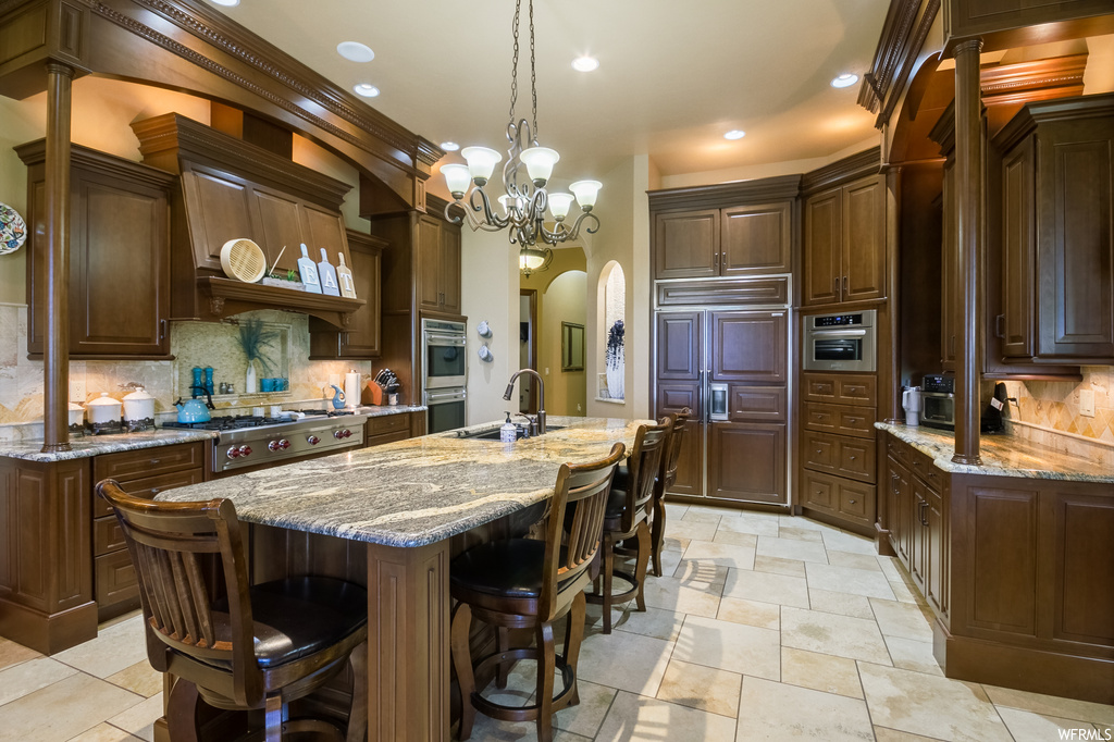 Kitchen with kitchen island with sink, backsplash, a chandelier, dark brown cabinets, custom exhaust hood, light tile floors, a center island, light stone countertops, and stainless steel appliances
