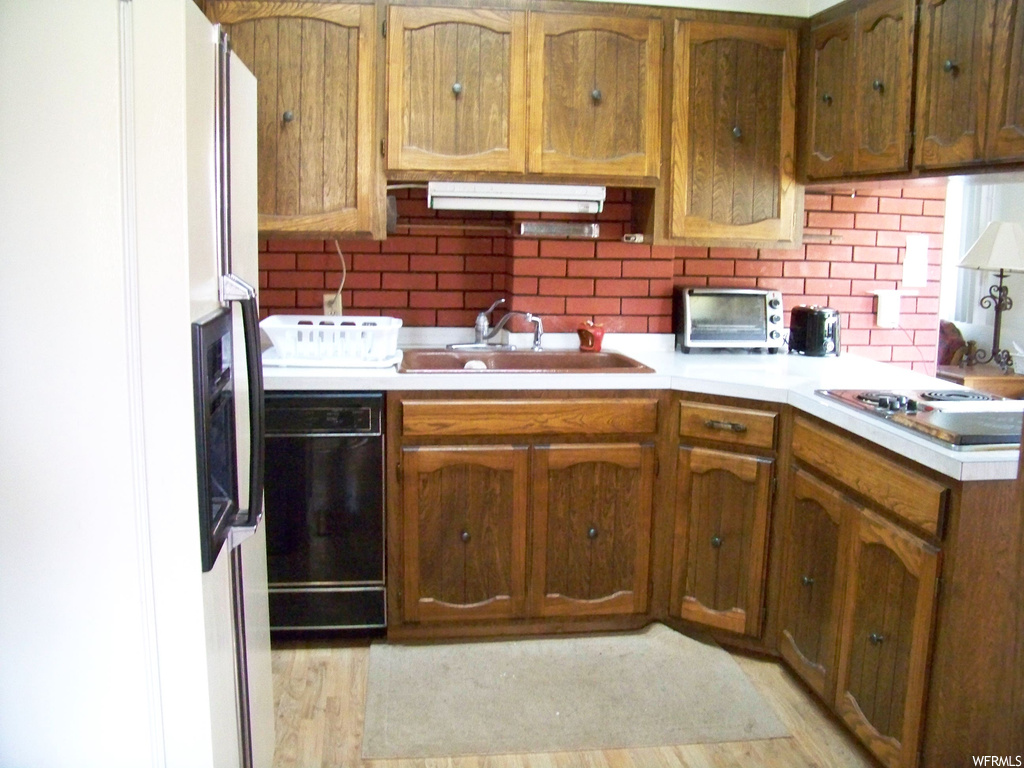 Kitchen with white refrigerator, backsplash, brown cabinets, light countertops, light hardwood floors, electric cooktop, and black dishwasher