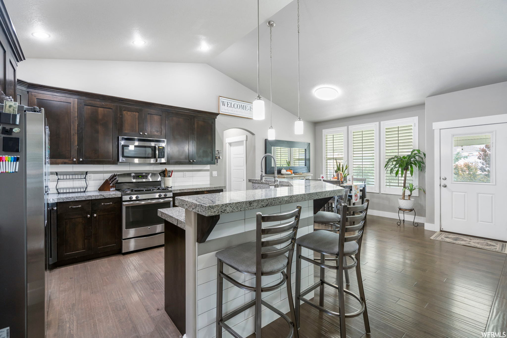 Kitchen featuring appliances with stainless steel finishes, a kitchen island, lofted ceiling, dark brown cabinetry, light countertops, hanging light fixtures, kitchen island with sink, backsplash, and light hardwood floors