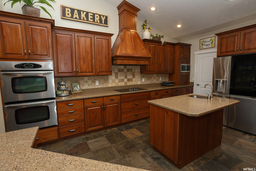 Kitchen with kitchen island with sink, backsplash, brown cabinets, appliances with stainless steel finishes, lofted ceiling, light stone countertops, dark tile floors, and custom range hood