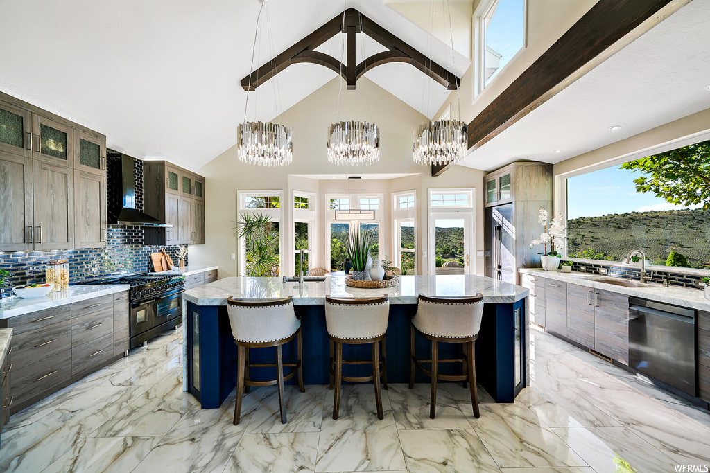 Kitchen with range with two ovens, a notable chandelier, a kitchen island, wall chimney exhaust hood, dishwasher, light stone countertops, kitchen island with sink, backsplash, a high ceiling, and light tile floors