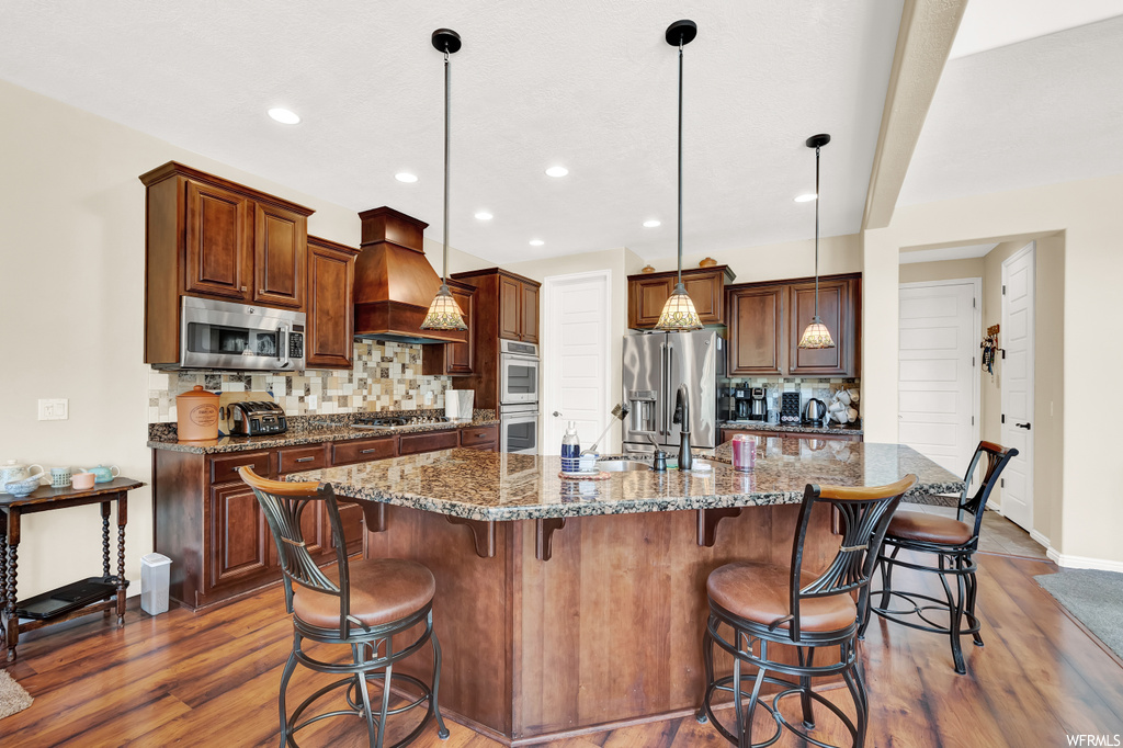 Kitchen with decorative light fixtures, backsplash, light hardwood floors, appliances with stainless steel finishes, premium range hood, a center island, and dark stone counters