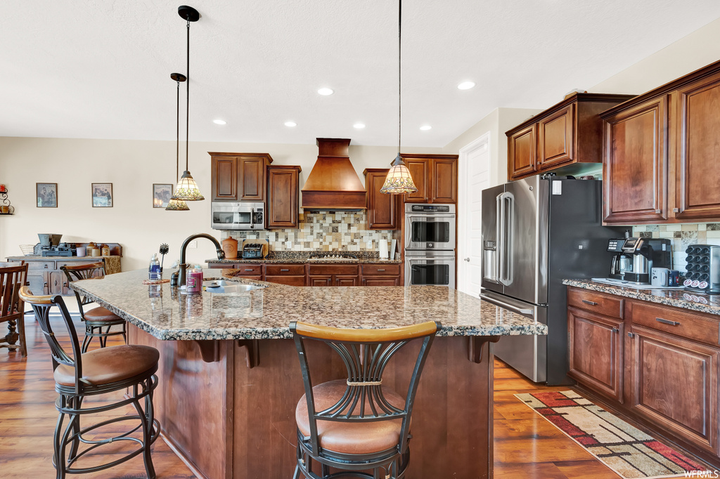 Kitchen with a kitchen island, custom range hood, pendant lighting, stainless steel appliances, light stone counters, wood-type flooring, and backsplash