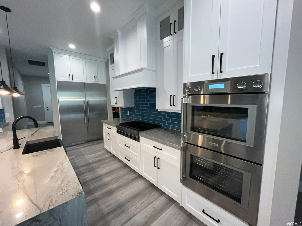 Kitchen featuring hanging light fixtures, backsplash, white cabinetry, hardwood flooring, stainless steel appliances, and custom range hood