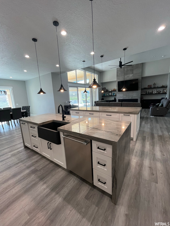 Kitchen featuring stainless steel dishwasher, plenty of natural light, white cabinetry, a textured ceiling, decorative light fixtures, light stone counters, hardwood floors, and kitchen island with sink