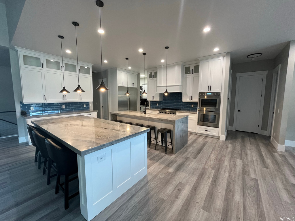 Kitchen with backsplash, light countertops, white cabinetry, a center island with sink, a center island, hardwood floors, pendant lighting, stainless steel double oven, and custom range hood