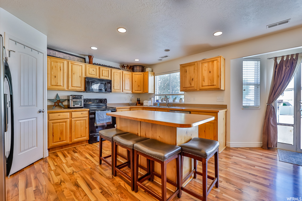 Kitchen featuring a textured ceiling, brown cabinets, black appliances, light hardwood floors, a center island, and plenty of natural light