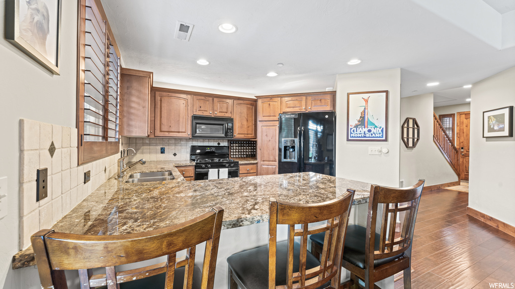 Kitchen with backsplash, brown cabinets, black appliances, a kitchen island, light hardwood flooring, and dark stone countertops