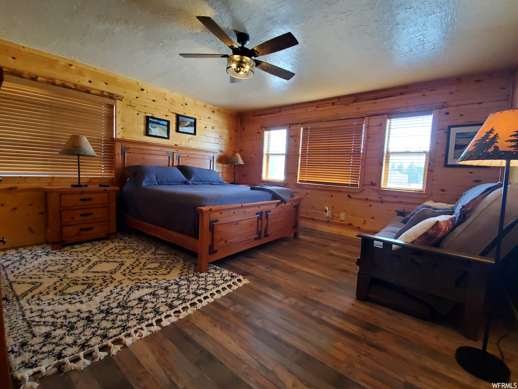 Hardwood floored bedroom with ceiling fan, wood walls, multiple windows, and a textured ceiling