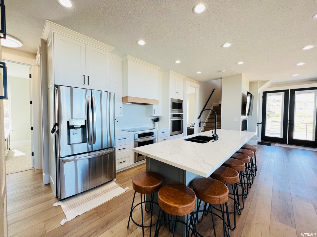 Kitchen with appliances with stainless steel finishes, a center island, a textured ceiling, an island with sink, white cabinets, light countertops, premium range hood, and light hardwood floors