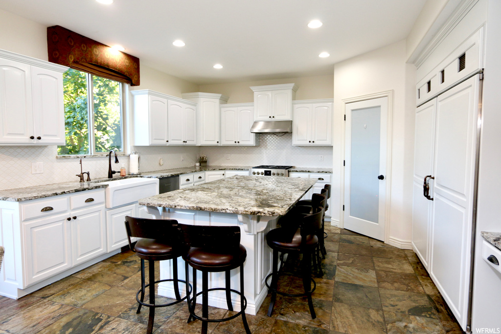 Kitchen featuring range, backsplash, a kitchen island, white cabinetry, dishwashing machine, light stone countertops, and dark tile floors