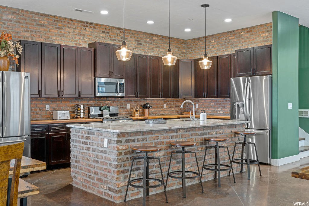 Kitchen with brick wall, stainless steel appliances, a center island, tile floors, pendant lighting, dark brown cabinetry, light stone countertops, and backsplash