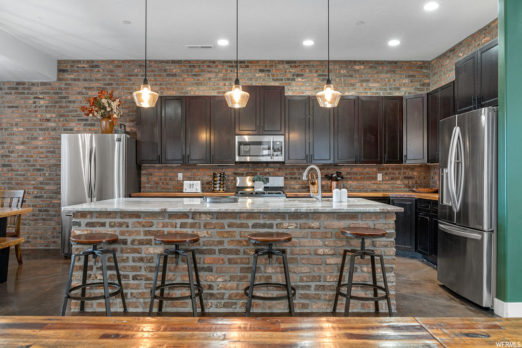 Kitchen with stone counters, decorative light fixtures, backsplash, brick wall, a kitchen island, dark brown cabinetry, and stainless steel appliances