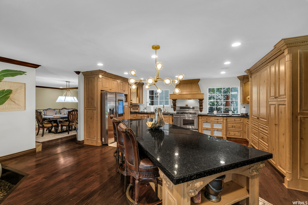 Kitchen with dark hardwood floors, brown cabinets, appliances with stainless steel finishes, a center island, crown molding, dark stone counters, and custom range hood