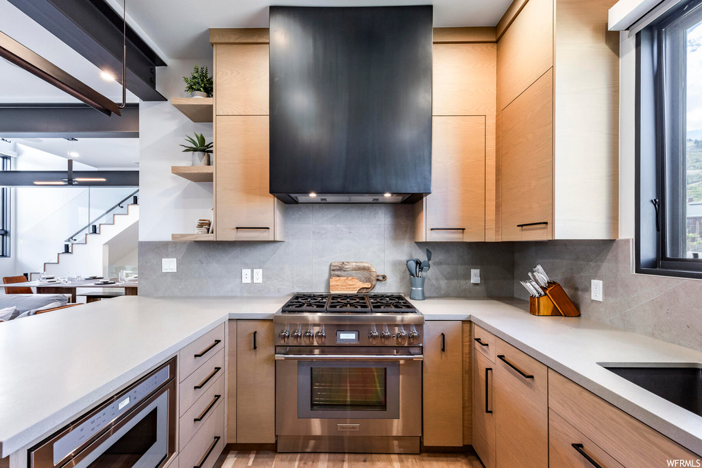 Kitchen with wall chimney range hood, high end stainless steel range oven, hardwood flooring, light countertops, light brown cabinetry, and backsplash