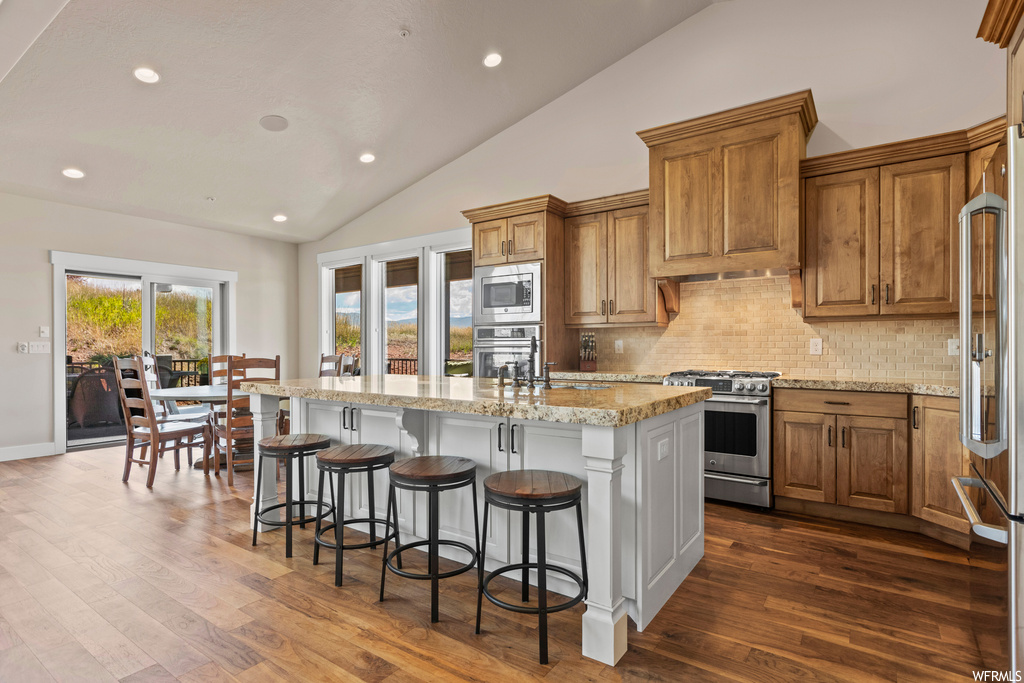 Kitchen featuring appliances with stainless steel finishes, a kitchen island, light stone countertops, vaulted ceiling, light hardwood flooring, and backsplash