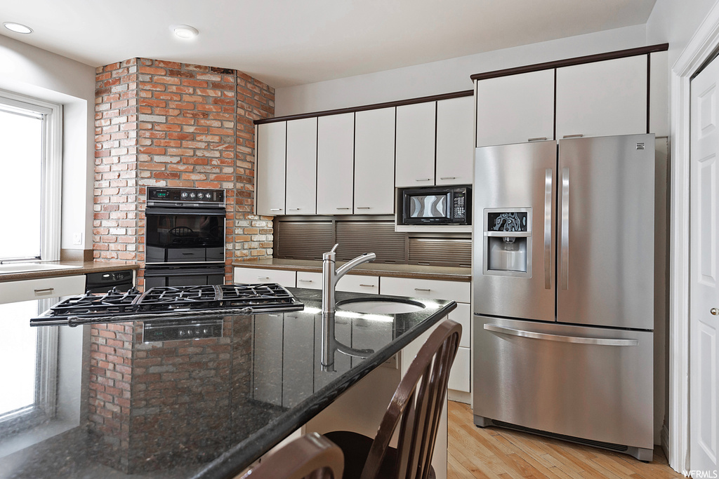 Kitchen featuring brick wall, black appliances, dark stone counters, white cabinets, and light hardwood floors