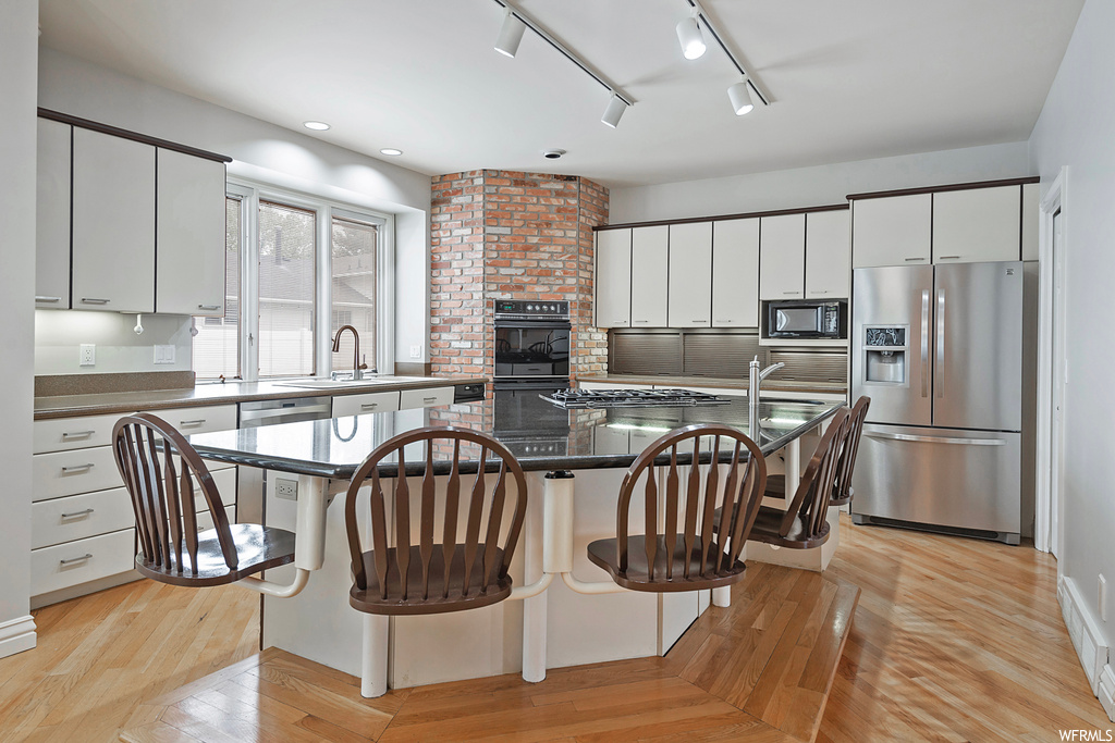 Kitchen featuring brick wall, rail lighting, a kitchen island, black appliances, dark countertops, white cabinets, and light hardwood floors