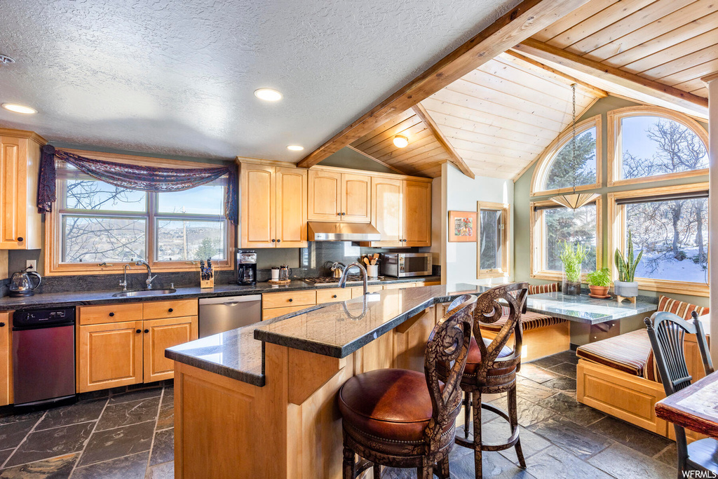 Kitchen featuring stainless steel appliances, a textured ceiling, a center island, tile flooring, dark countertops, a wealth of natural light, brown cabinets, and vaulted ceiling with beams