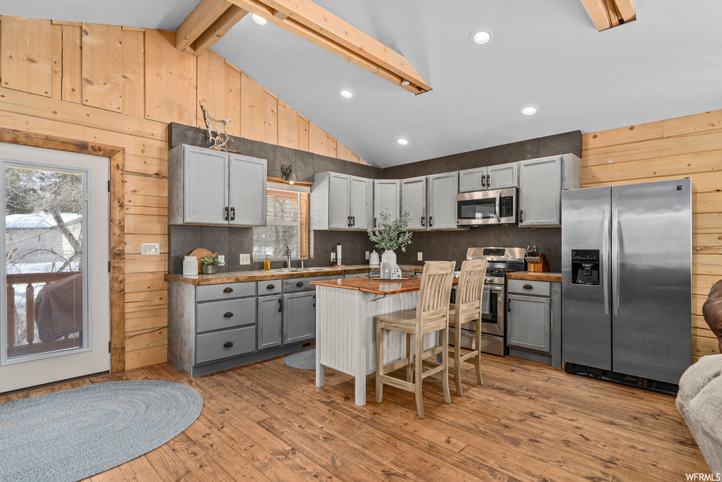 Kitchen with a kitchen island, stainless steel appliances, white cabinets, wood walls, lofted ceiling with beams, backsplash, light hardwood floors, and a high ceiling