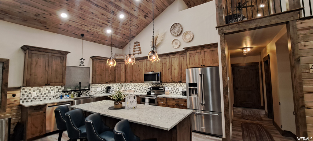 Kitchen with appliances with stainless steel finishes, a center island, decorative light fixtures, wooden ceiling, lofted ceiling, light stone countertops, hardwood flooring, dark brown cabinets, a center island with sink, backsplash, and a high ceiling