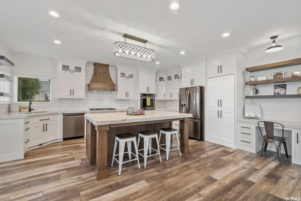 Kitchen featuring appliances with stainless steel finishes, a kitchen island, white cabinets, custom exhaust hood, wood-type flooring, light countertops, and backsplash