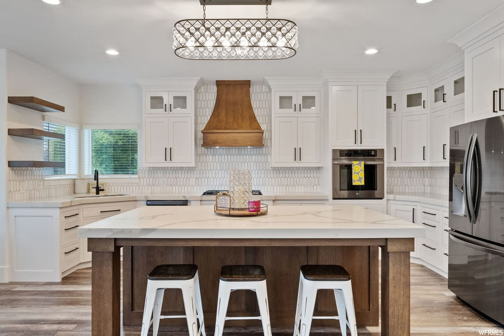 Kitchen with backsplash, light countertops, white cabinetry, appliances with stainless steel finishes, light hardwood flooring, a center island, premium range hood, pendant lighting, and a kitchen island with sink
