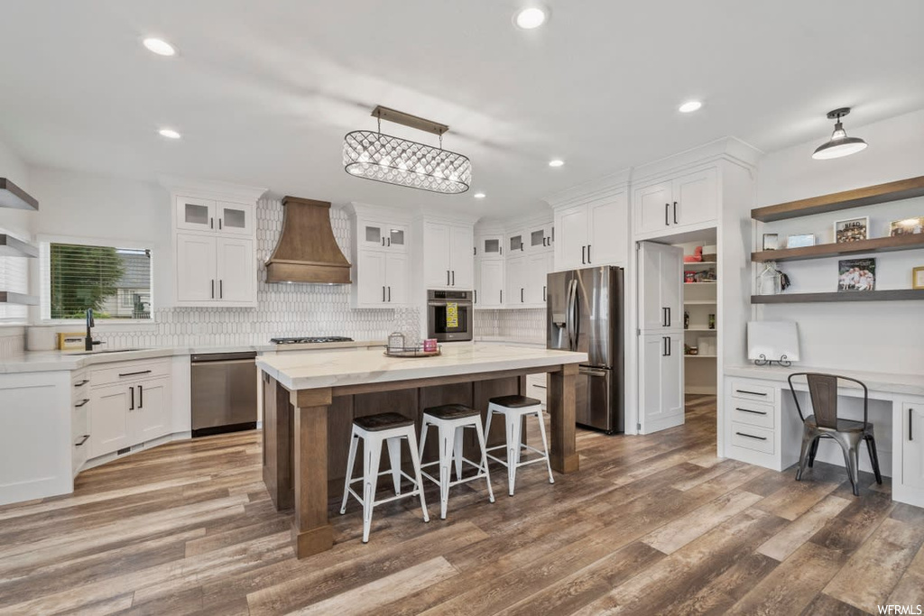 Kitchen featuring a kitchen island, white cabinetry, stainless steel appliances, hardwood flooring, light countertops, premium range hood, and backsplash