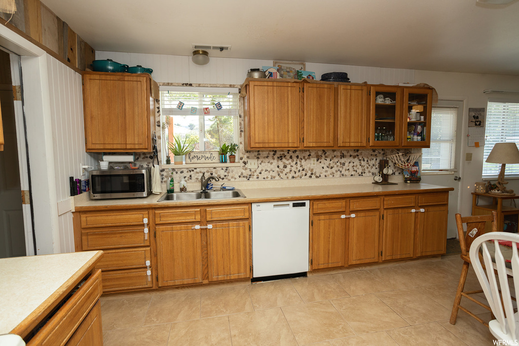 Kitchen with backsplash, brown cabinets, light countertops, white dishwasher, and light tile flooring