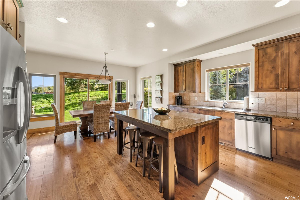 Kitchen featuring stone counters, a textured ceiling, backsplash, a kitchen island, light hardwood flooring, plenty of natural light, a kitchen island with sink, and stainless steel appliances
